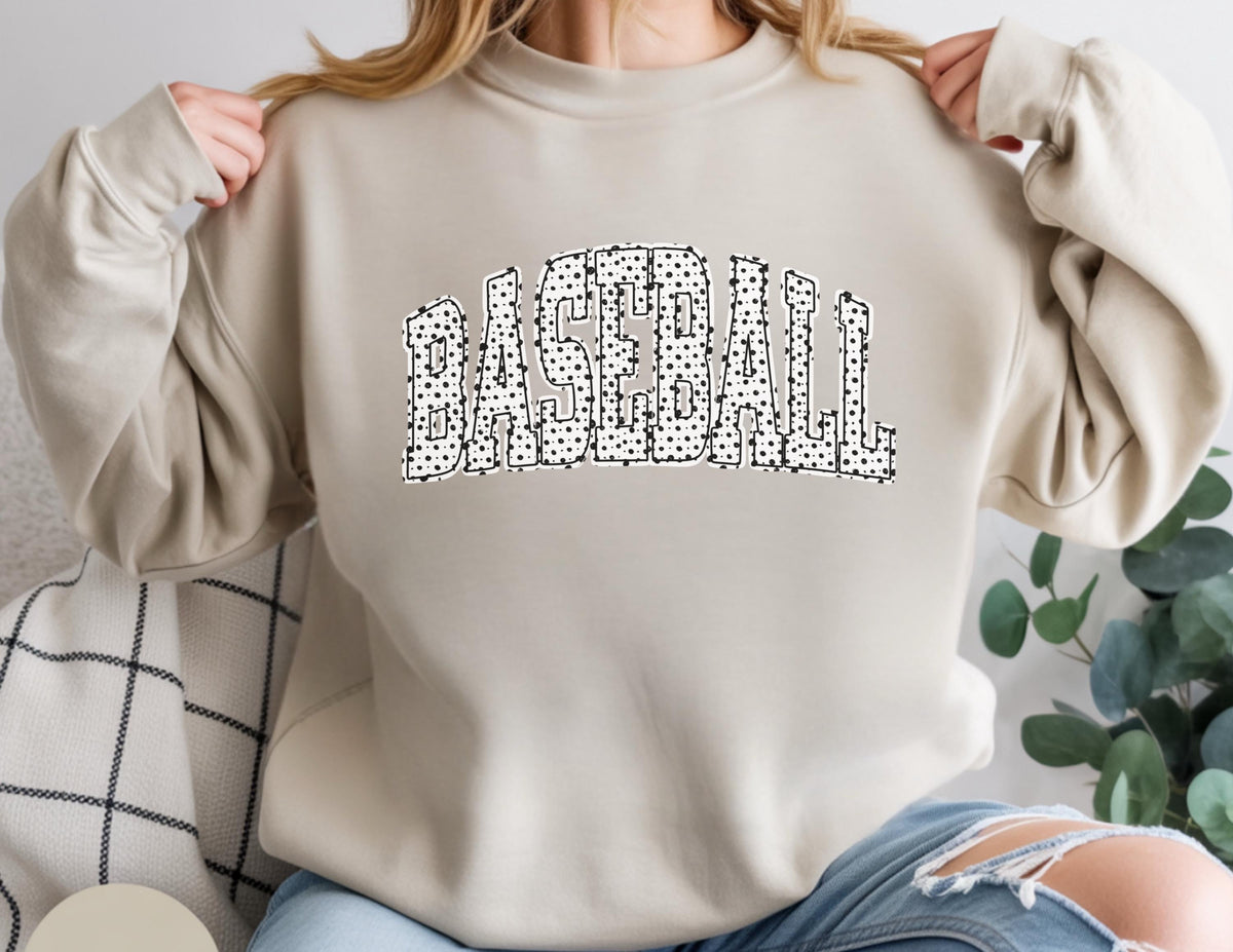 May include: A beige sweatshirt with the word "BASEBALL" in large, black and white polka dot letters across the chest. The sweatshirt has long sleeves and a crew neck.