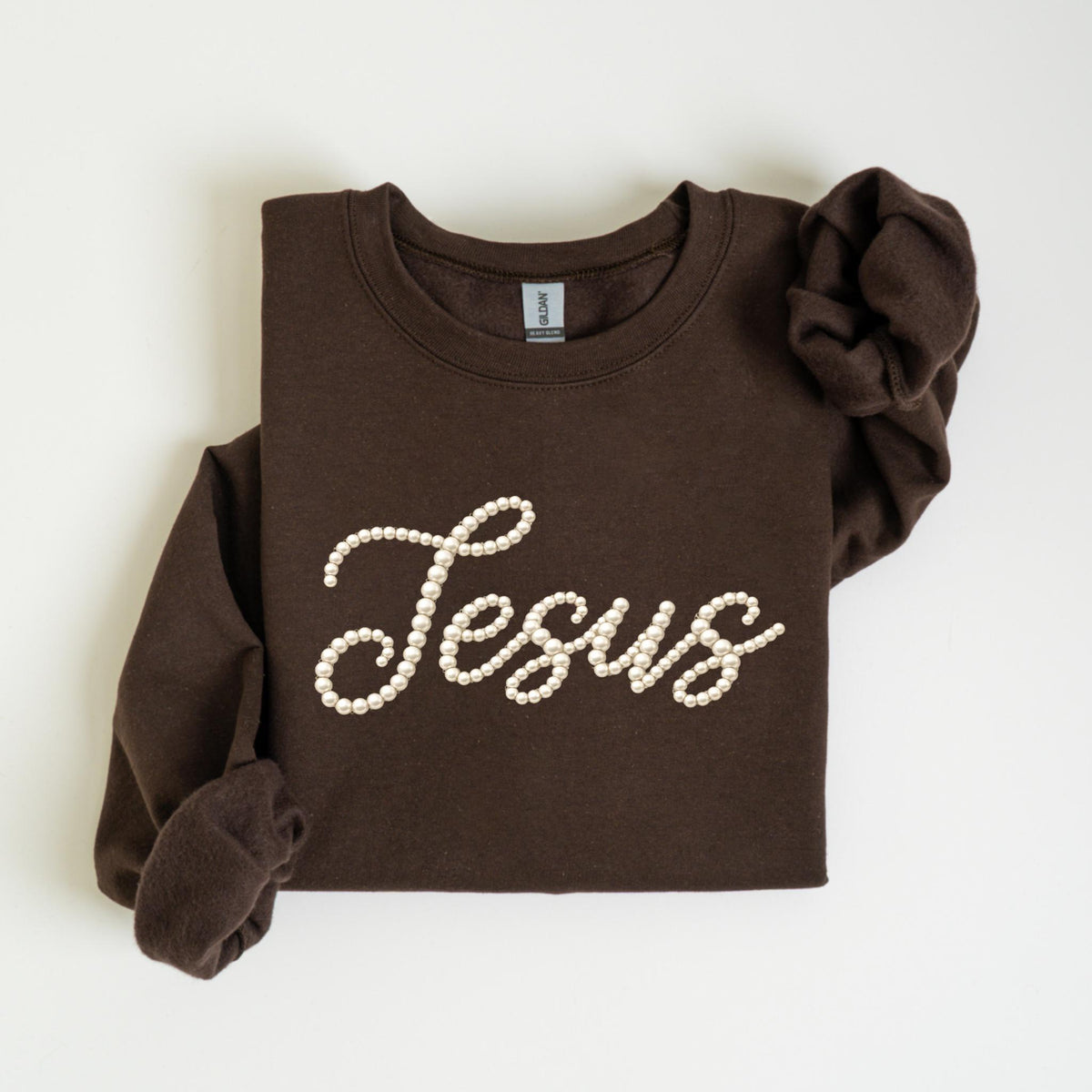 May include: A dark brown sweatshirt with the word "Jesus" spelled out in white pearl-like beads. The long-sleeved top is folded neatly on a white surface.
