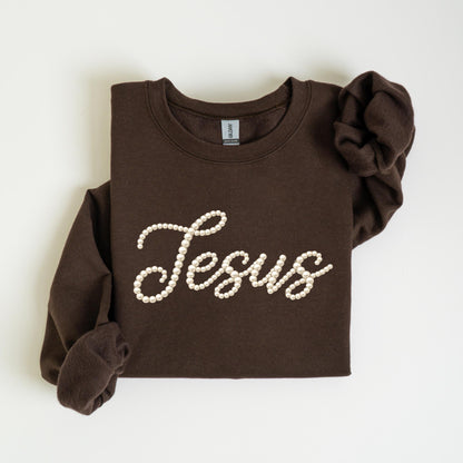 May include: A dark brown sweatshirt with the word "Jesus" spelled out in white pearl-like beads. The long-sleeved top is folded neatly on a white surface.
