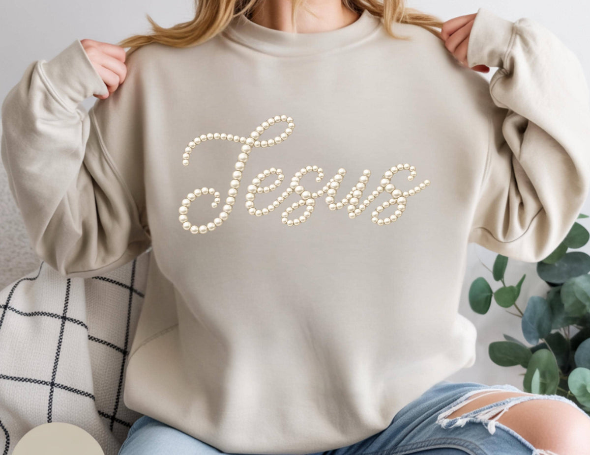 May include: A beige sweatshirt with the word "Jesus" spelled out in a cursive font using faux pearls. The sweatshirt has long sleeves and a crew neck. The person in the image is wearing blue jeans.