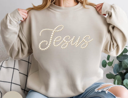 May include: A beige sweatshirt with the word "Jesus" spelled out in a cursive font using faux pearls. The sweatshirt has long sleeves and a crew neck. The person in the image is wearing blue jeans.