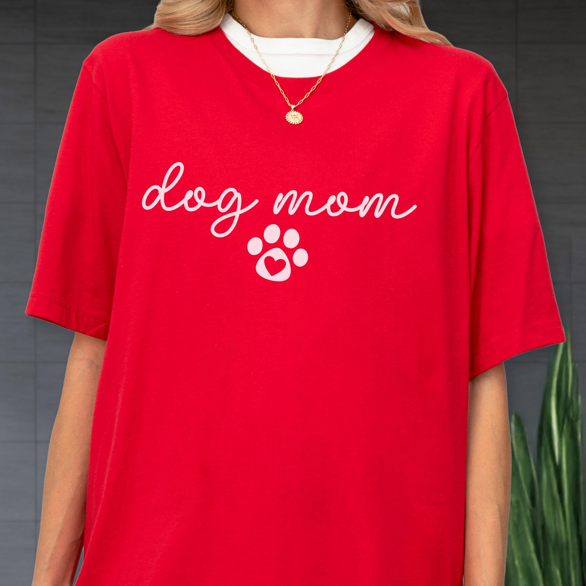 May include: A red t-shirt with the words "dog mom" in white script, along with a white paw print with a heart in the center. The shirt has a white collar and is worn with a gold necklace.