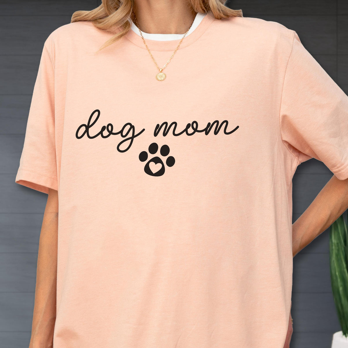 May include: A light peach-colored t-shirt with the words "dog mom" in a cursive font and a paw print with a heart in the center. The shirt has a white collar and short sleeves.