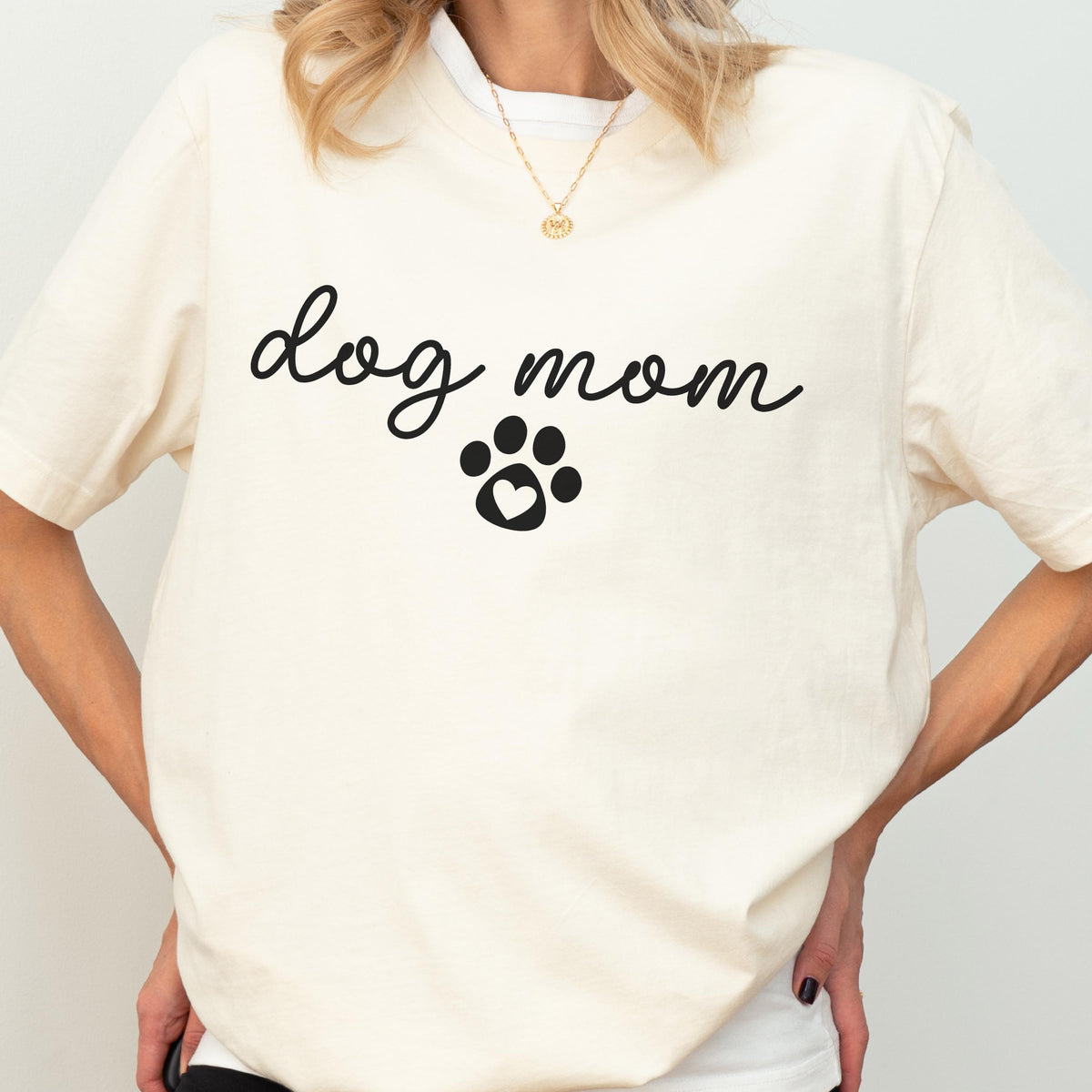 May include: Cream-colored t-shirt with the words "dog mom" in a cursive font, and a paw print with a heart in the center. The shirt has short sleeves and a crew neck.