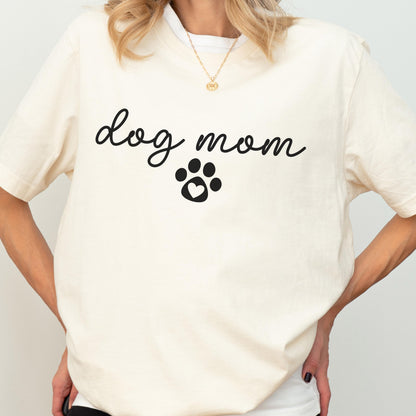 May include: Cream-colored t-shirt with the words "dog mom" in a cursive font, and a paw print with a heart in the center. The shirt has short sleeves and a crew neck.