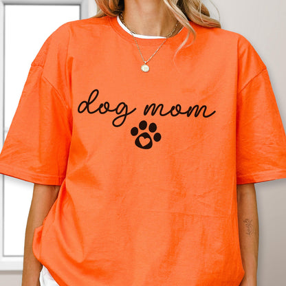 May include: Bright orange t-shirt with the words "dog mom" in a cursive font, and a black paw print with a heart in the center. The shirt has a relaxed fit and short sleeves. The model is wearing a gold necklace.