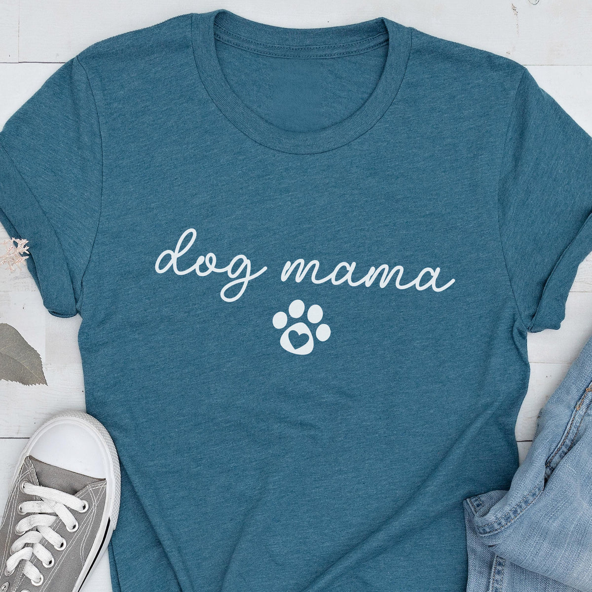 May include: Teal-colored t-shirt with the words "dog mama" in a cursive font and a white paw print graphic. The shirt is made of a soft material and is suitable for casual wear. A pair of gray sneakers and blue jeans are visible.