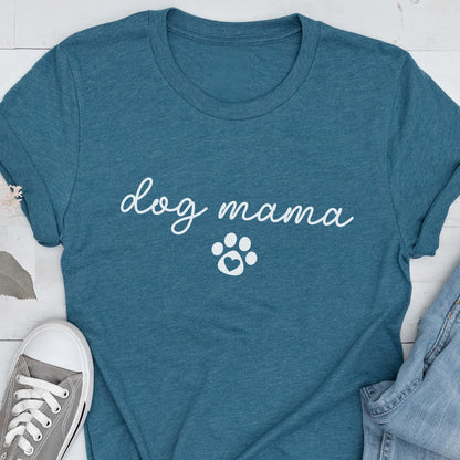 May include: Teal-colored t-shirt with the words "dog mama" in a cursive font and a white paw print graphic. The shirt is made of a soft material and is suitable for casual wear. A pair of gray sneakers and blue jeans are visible.