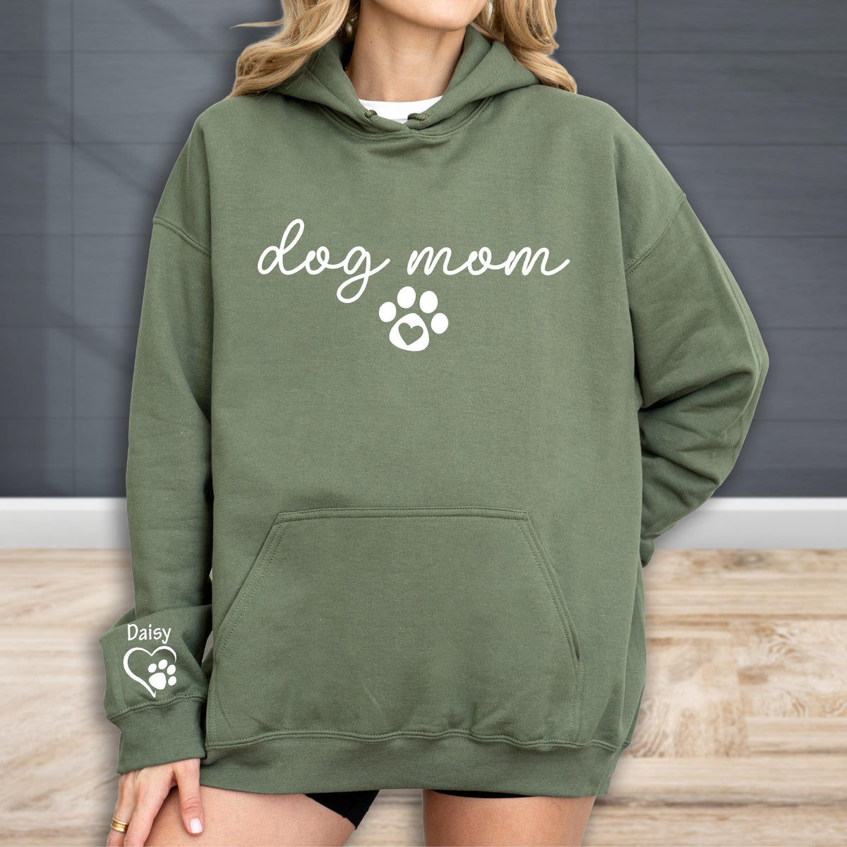 May include: An olive green hoodie with the words "dog mom" in white script, along with a paw print design. The hoodie has a front pocket and a drawstring hood. The left sleeve has a smaller paw print design with the name "Daisy".