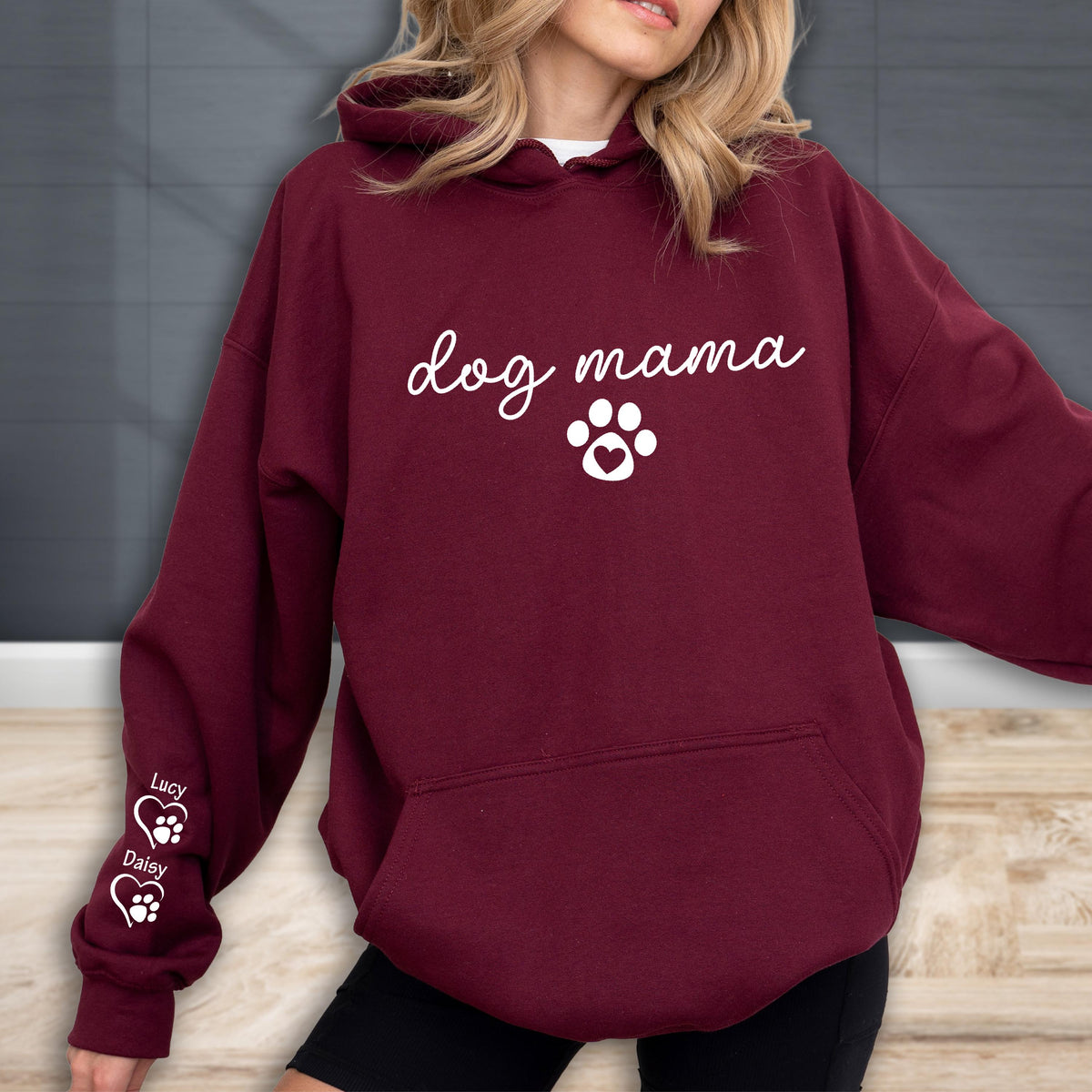May include: Burgundy hoodie with the words "dog mama" in white script, a paw print with a heart, and the names "Lucy" and "Daisy" with paw prints on the sleeve. The hoodie has a drawstring hood and a kangaroo pocket.