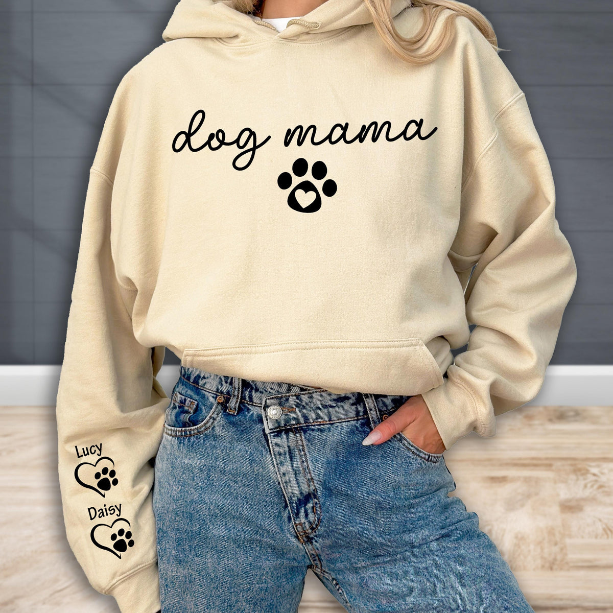 May include: A beige hoodie with the words "dog mama" in a script font, along with a paw print design with a heart. The left sleeve has the names "Lucy" and "Daisy" with paw print and heart designs. The person is wearing blue jeans.