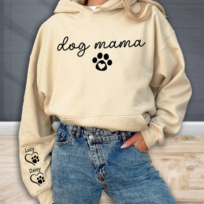 May include: A beige hoodie with the words "dog mama" in a script font, along with a paw print design with a heart. The left sleeve has the names "Lucy" and "Daisy" with paw print and heart designs. The person is wearing blue jeans.