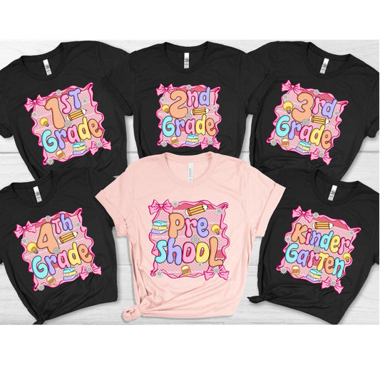 May include: A collection of black and pink t-shirts with school-themed graphics. Each shirt features a grade level, such as "1st Grade" or "Kindergarten," in colorful bubble letters, surrounded by bows, pencils, and books.