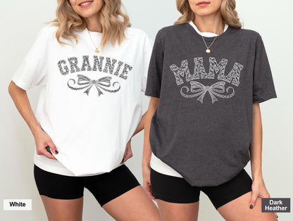 May include: Two women wearing white and dark heather gray t-shirts. The white shirt says "GRANNIE" in a decorative font above a bow design. The dark heather gray shirt says "MAMA" in a decorative font above a bow design. Both shirts have short sleeves.