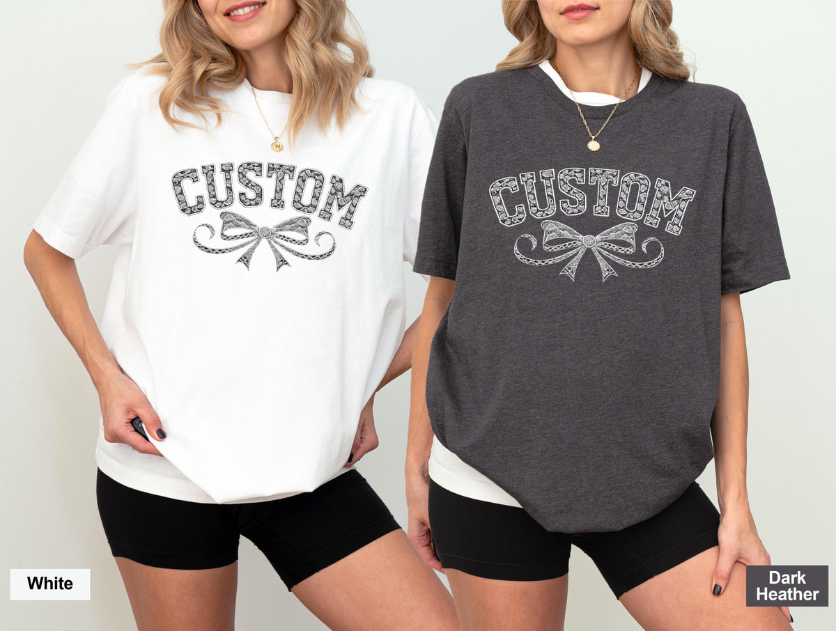 May include: Two t-shirts, one white and one dark heather, each featuring the word "CUSTOM" above a decorative bow design. The shirts are short-sleeved and worn with black shorts. The white shirt has a white background, and the dark heather shirt has a dark background.