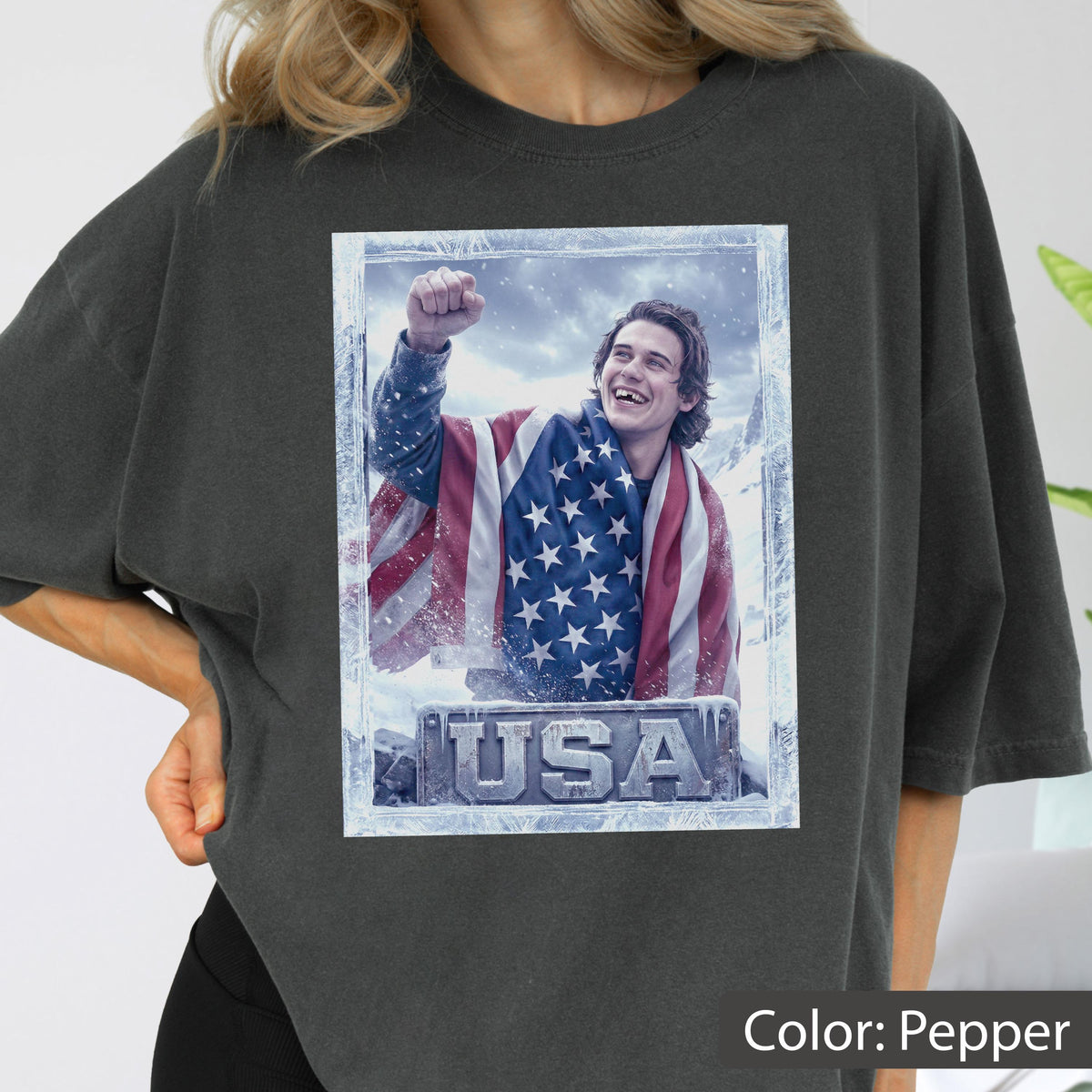 May include: Pepper-colored t-shirt featuring a graphic of a person wrapped in an American flag, with the word "USA" below. The image has a frosted border. The shirt is a relaxed fit.