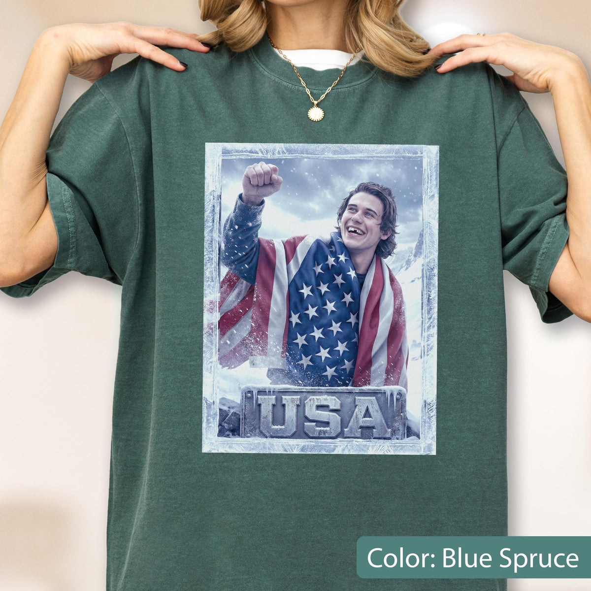 May include: Blue spruce t-shirt featuring a graphic of a person wrapped in an American flag, with the word "USA" at the bottom. The graphic is framed in a frosty border. The shirt has a crew neck and short sleeves.