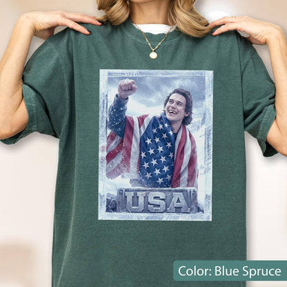 May include: Blue spruce t-shirt featuring a graphic of a person wrapped in an American flag, with the word "USA" at the bottom. The graphic is framed in a frosty border. The shirt has a crew neck and short sleeves.