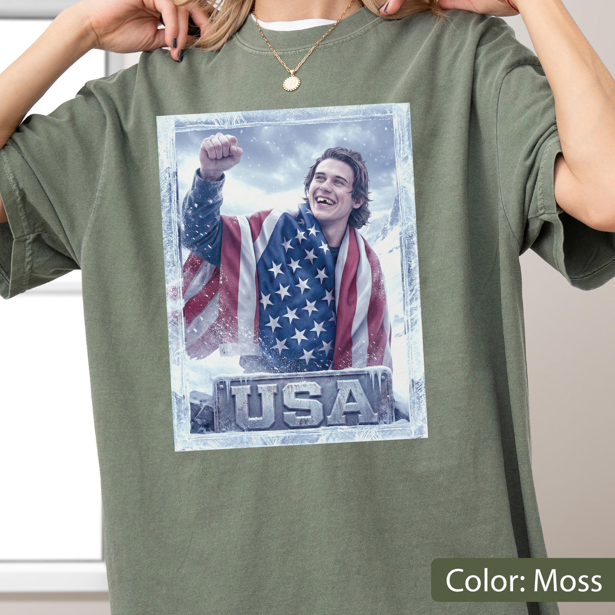 May include: Moss green t-shirt featuring a graphic of a person wrapped in an American flag, with the word "USA" at the bottom. The graphic is framed with a frosty border. The shirt is a solid color.