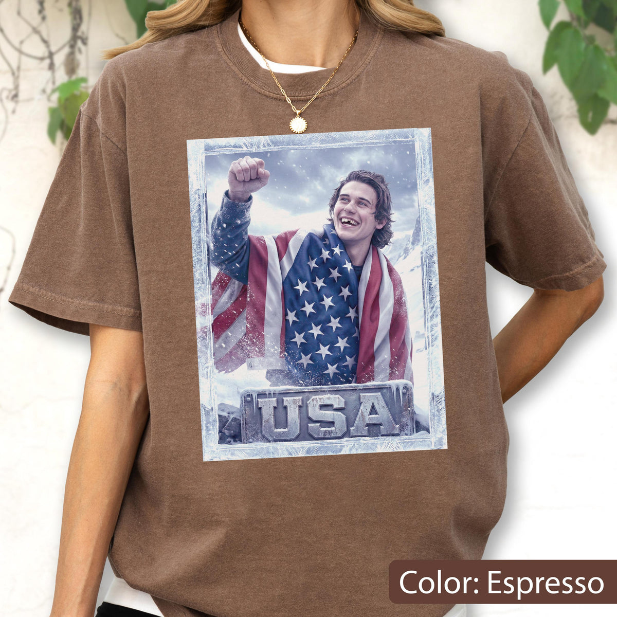 May include: Espresso-colored t-shirt featuring a framed image of a person wrapped in an American flag, with the word "USA" at the bottom. The person is smiling and has their fist raised. The shirt has a crew neck.