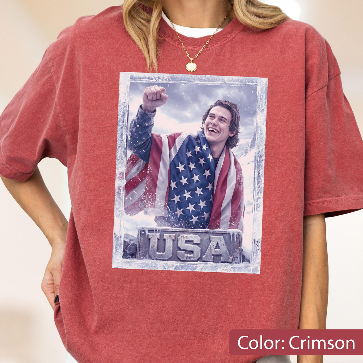 May include: Crimson-colored t-shirt featuring a framed image of a person draped in an American flag, with the word "USA" at the bottom. The person is smiling and has their fist raised in the air. The shirt is a solid color.