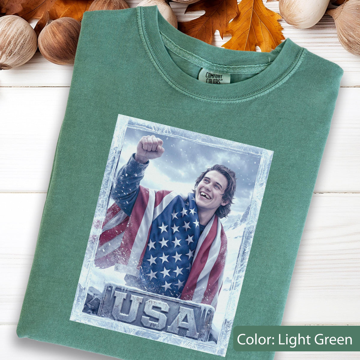 May include: Light green t-shirt featuring a graphic of a person wrapped in an American flag, with the word "USA" at the bottom. The shirt is a solid light green color. The tag on the shirt reads "Comfort Colors".