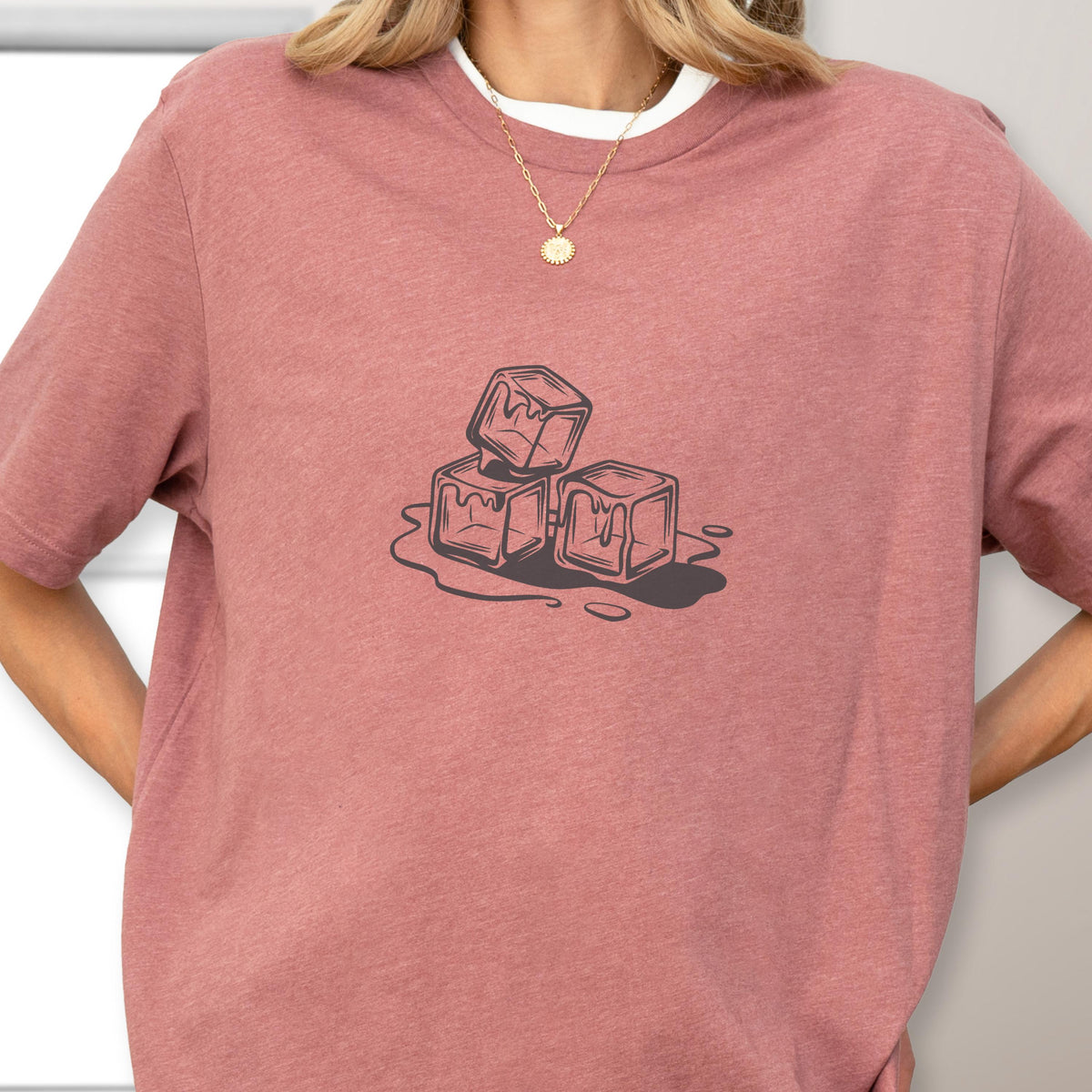 May include: A dusty rose t-shirt with a white crew neck collar. The shirt features a graphic of three melting ice cubes in a dark gray color. The ice cubes are stacked on top of each other.