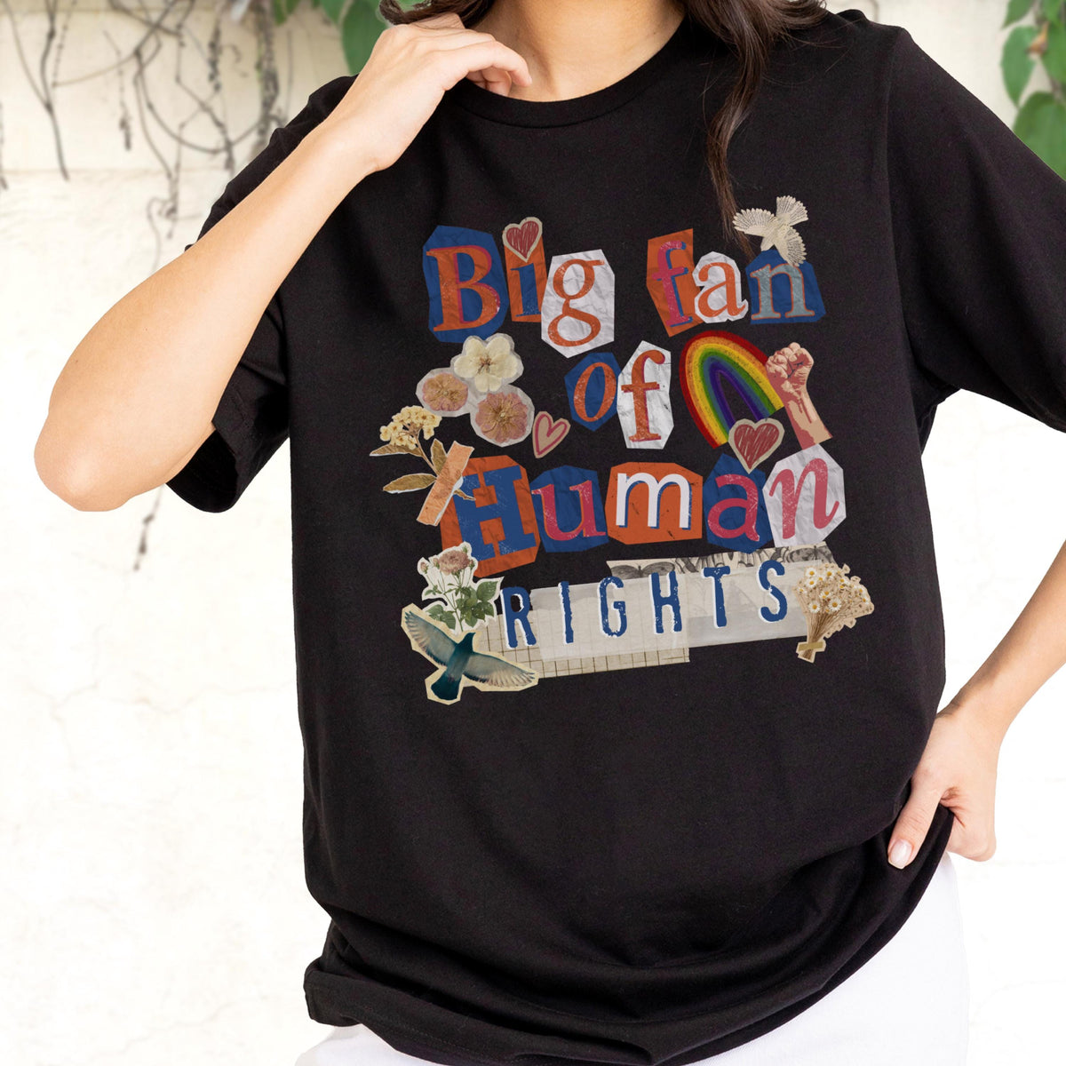 May include: Black t-shirt with a colorful collage design. The design includes the words "Big Fan of Human Rights" in a mix of fonts and colors, along with images of flowers, a rainbow, and a raised fist.