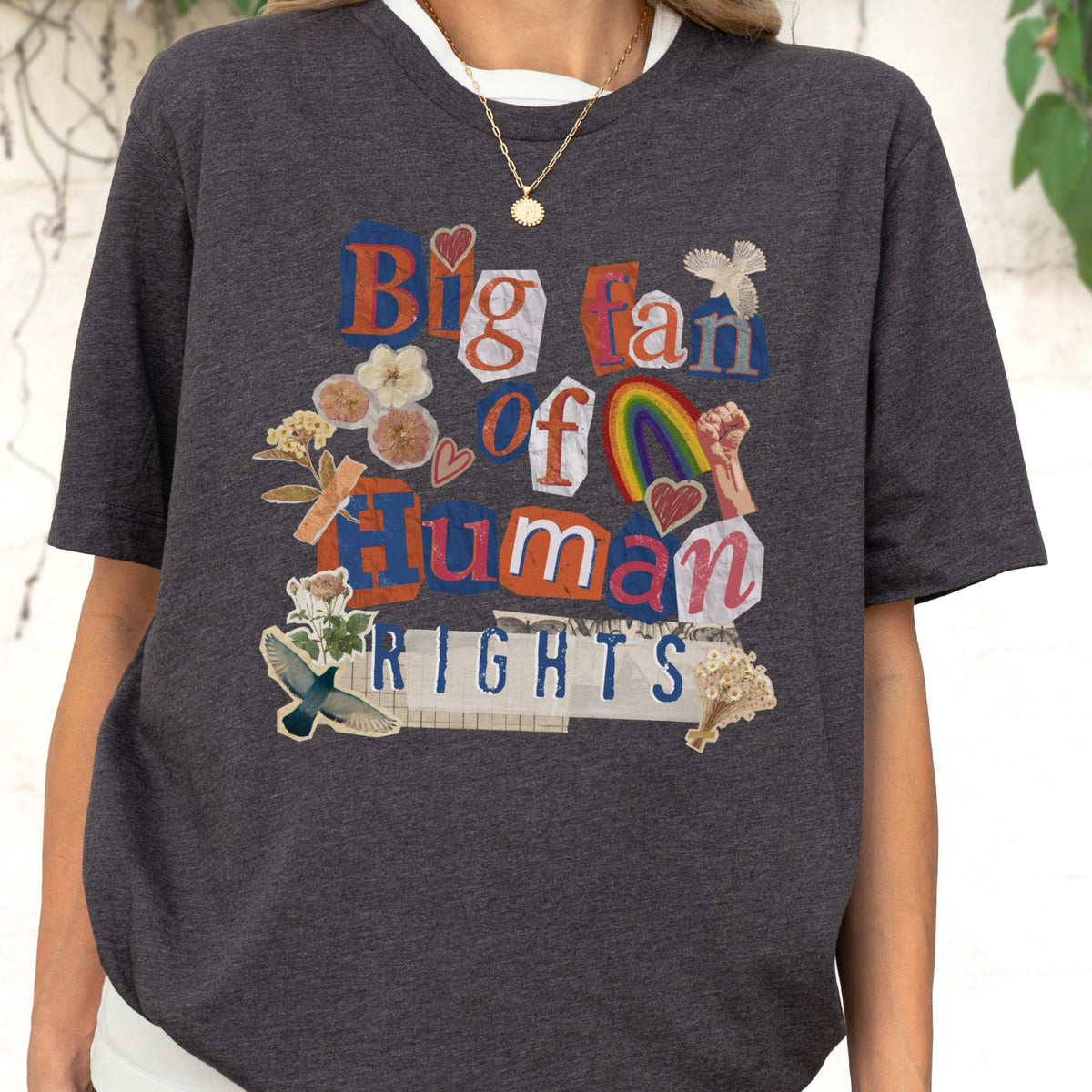 May include: Dark gray t-shirt with a white collar featuring a collage design. The design includes the text "Big fan of Human Rights" in a mix of fonts and colors, along with floral and rainbow graphics. A gold necklace is visible.