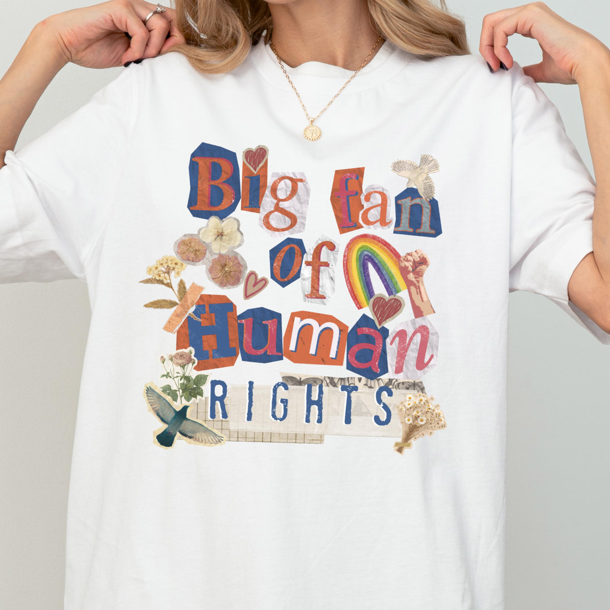 May include: White t-shirt with a colorful collage design. The design features the text "Big fan of Human Rights" in a mix of fonts and colors, along with illustrations of a rainbow, flowers, and a raised fist. The shirt is a casual, everyday wear item.