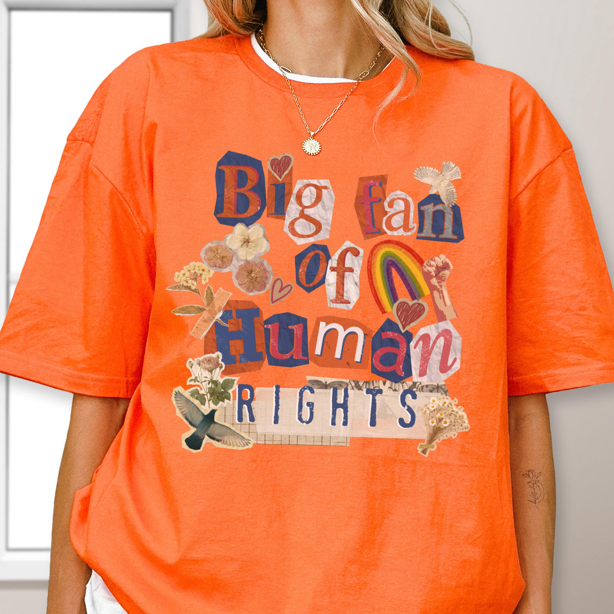 May include: An orange t-shirt with the text "Big fan of Human Rights" in a collage style. The design includes a rainbow, flowers, and a bird. The shirt has a crew neck and short sleeves.