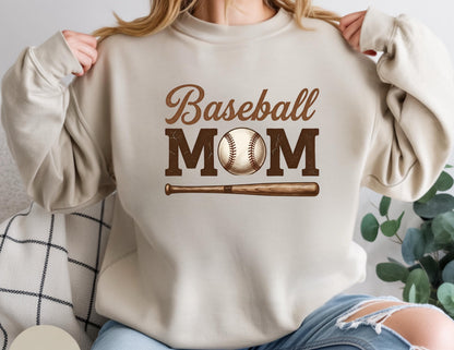May include: Beige sweatshirt with the words "Baseball MOM" in brown lettering. A baseball and bat graphic is centered below the text. The sweatshirt is a neutral color, suitable for casual wear.