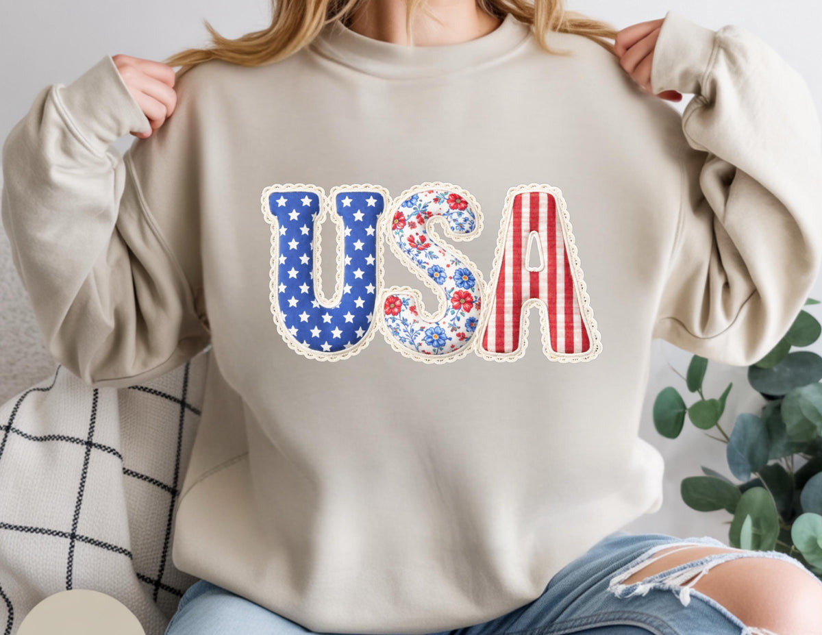 May include: Beige sweatshirt with the word "USA" in a patriotic design. The "U" is blue with white stars, the "S" has a floral pattern, and the "A" has red and white stripes. The letters are outlined with a scalloped trim.