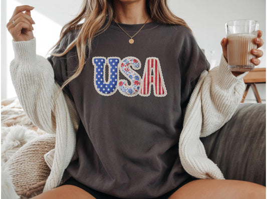 May include: Dark gray sweatshirt with the word "USA" in a patriotic design. The "U" is blue with white stars, the "S" has a floral pattern, and the "A" has red and white stripes. The sweatshirt is part of a casual clothing collection.