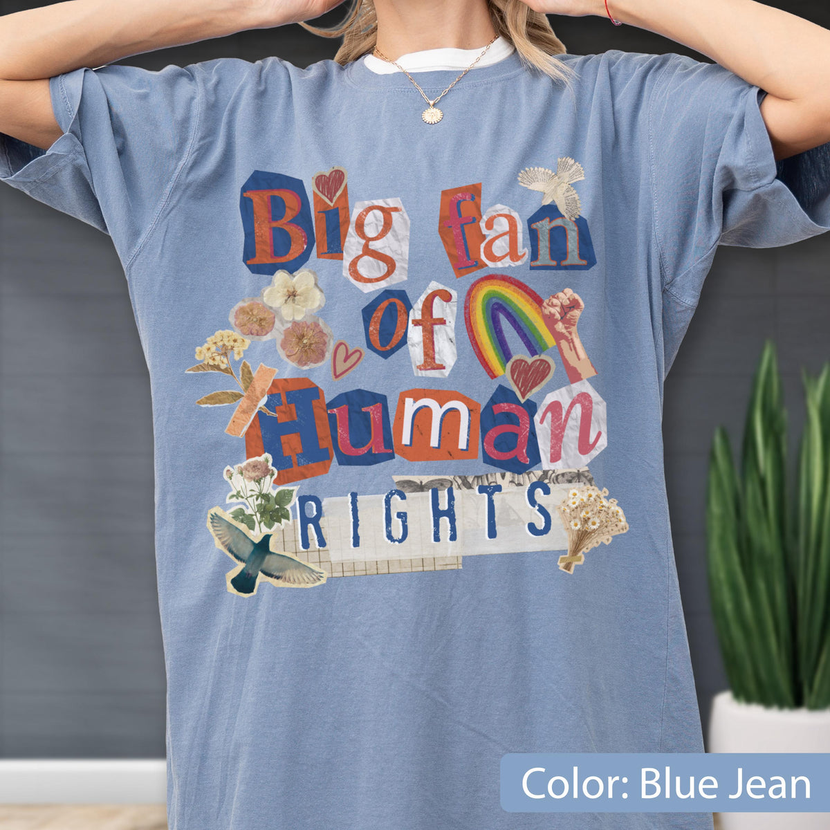 May include: Blue jean-colored t-shirt with the text "Big fan of Human Rights" in a collage style. The design includes a rainbow, hearts, and floral elements. The shirt has short sleeves and a crew neck.