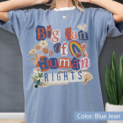May include: Blue jean-colored t-shirt with the text "Big fan of Human Rights" in a collage style. The design includes a rainbow, hearts, and floral elements. The shirt has short sleeves and a crew neck.