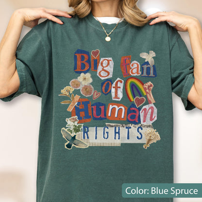 May include: Blue spruce-colored t-shirt with a collage design. The design includes the text "Big fan of Human Rights" in various fonts and colors, along with floral and rainbow graphics. The shirt has a crew neck and short sleeves.