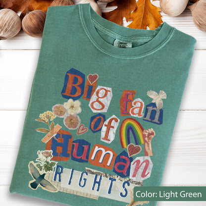 May include: Light green t-shirt with the text "Big fan of Human Rights" in a collage style. The design includes a rainbow, flowers, and a raised fist. The shirt is made of a soft material.