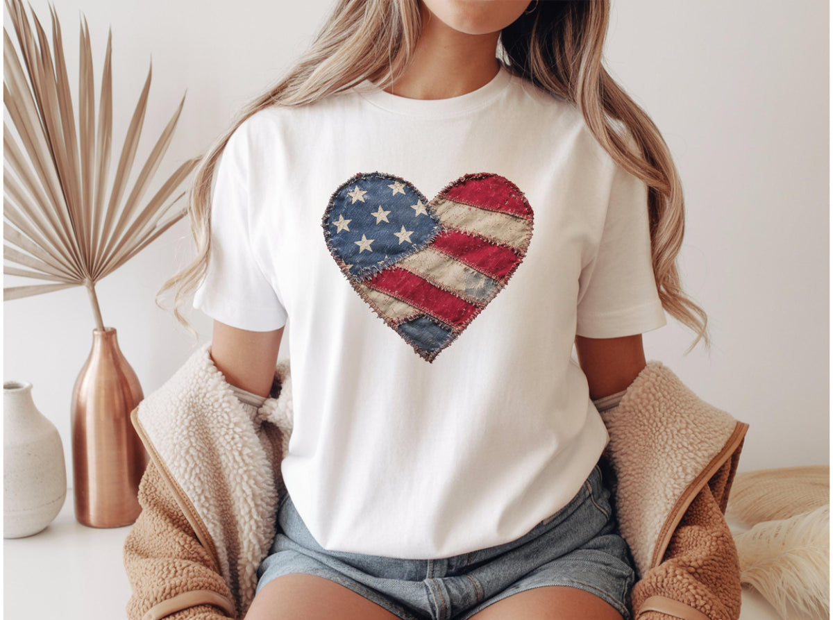 May include: White t-shirt featuring a heart-shaped design resembling the American flag. The heart is composed of blue fabric with white stars, and red and white striped fabric. The shirt is a casual style.