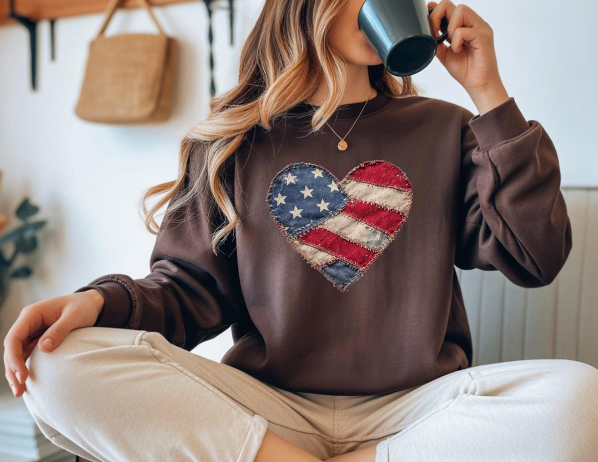 May include: A brown sweatshirt featuring a heart-shaped design resembling the American flag. The heart is made of blue, red, and white fabric. The sweatshirt has long sleeves and a crew neck. The overall style is casual and patriotic.