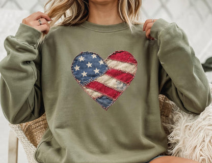 May include: Olive green sweatshirt featuring a heart-shaped patch with a distressed American flag design. The patch includes blue stars and red and white stripes. The sweatshirt has long sleeves and a crew neck.