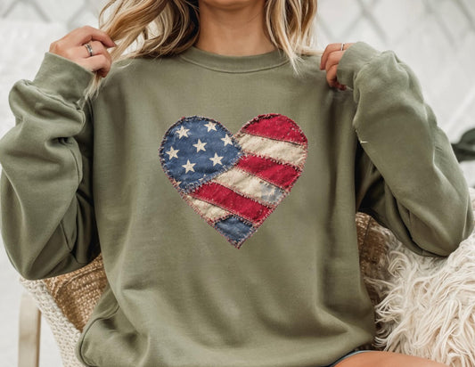 May include: Olive green sweatshirt featuring a heart-shaped patch with a distressed American flag design. The patch includes blue stars and red and white stripes. The sweatshirt has long sleeves and a crew neck.