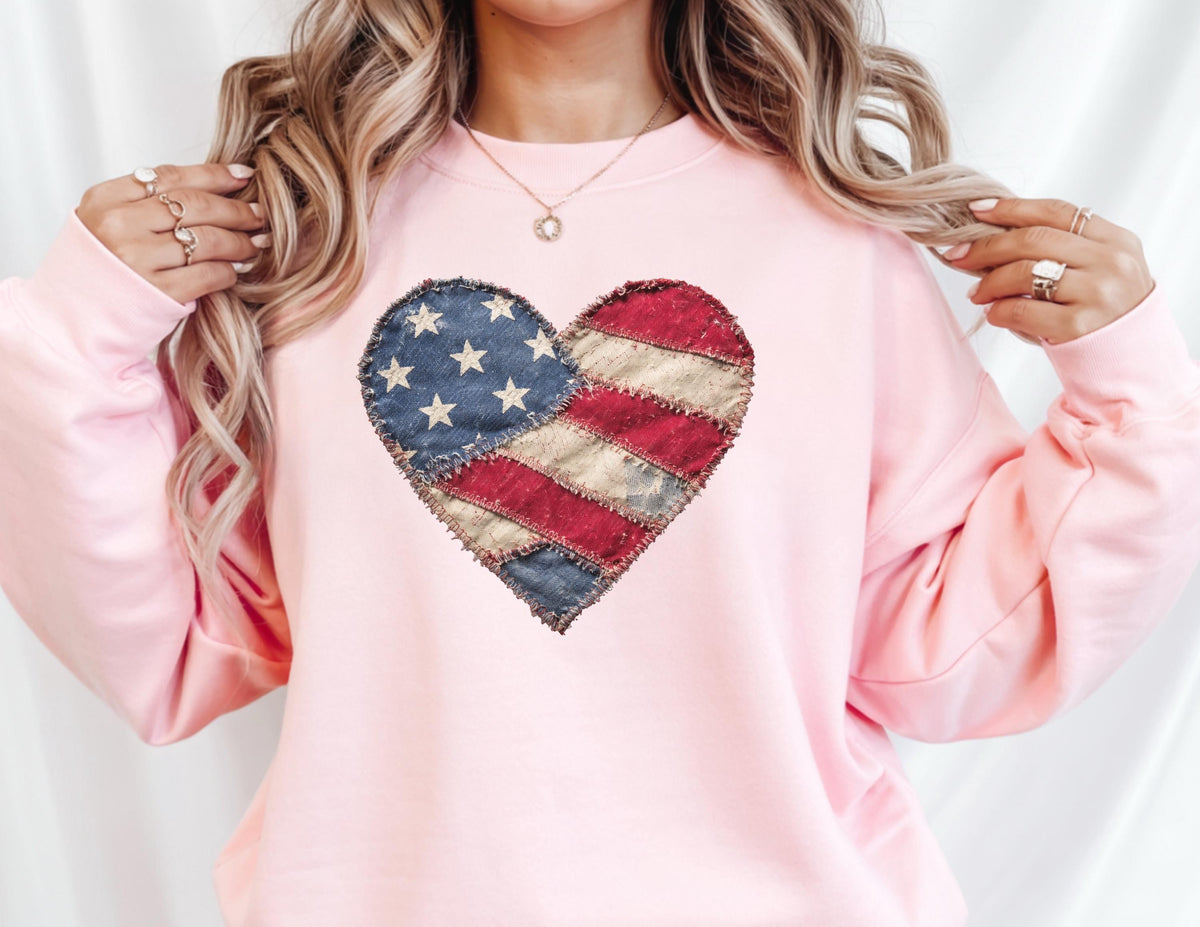 May include: A light pink sweatshirt featuring a heart-shaped design resembling the American flag. The heart is composed of blue fabric with white stars and red and white striped fabric, stitched together. The sweatshirt has long sleeves.