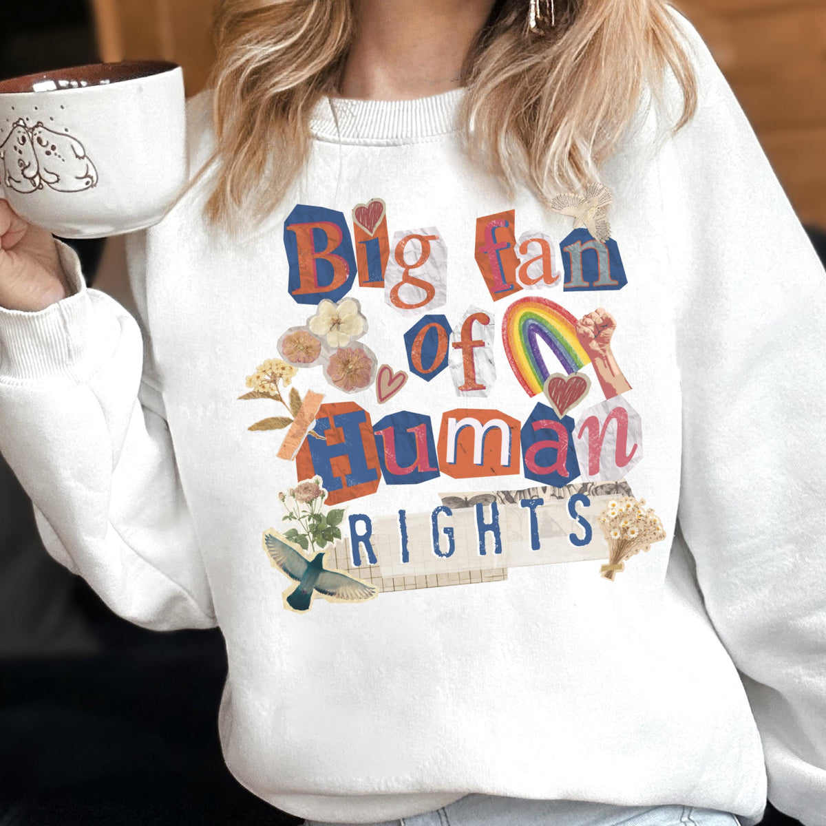 May include: White sweatshirt with the text "Big fan of Human Rights" in a collage style. The design includes a rainbow, flowers, and a bird. The sweatshirt is a crew neck style.