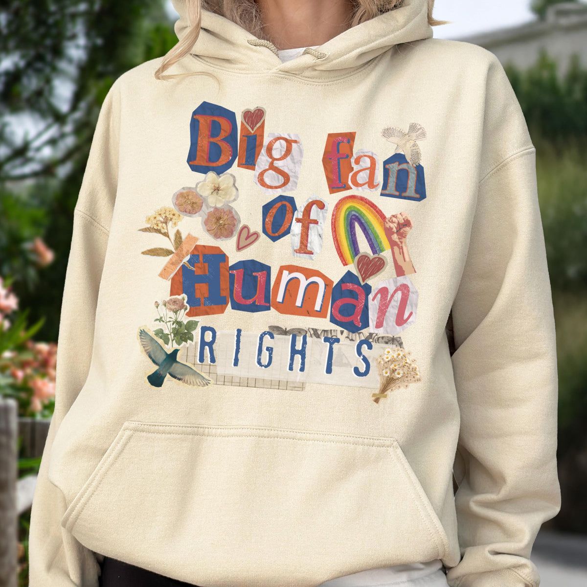 May include: Cream-colored hoodie with a graphic design that reads "Big Fan of Human Rights." The design incorporates a rainbow, hearts, flowers, and a bird. The text is in a collage style with various fonts and colors.