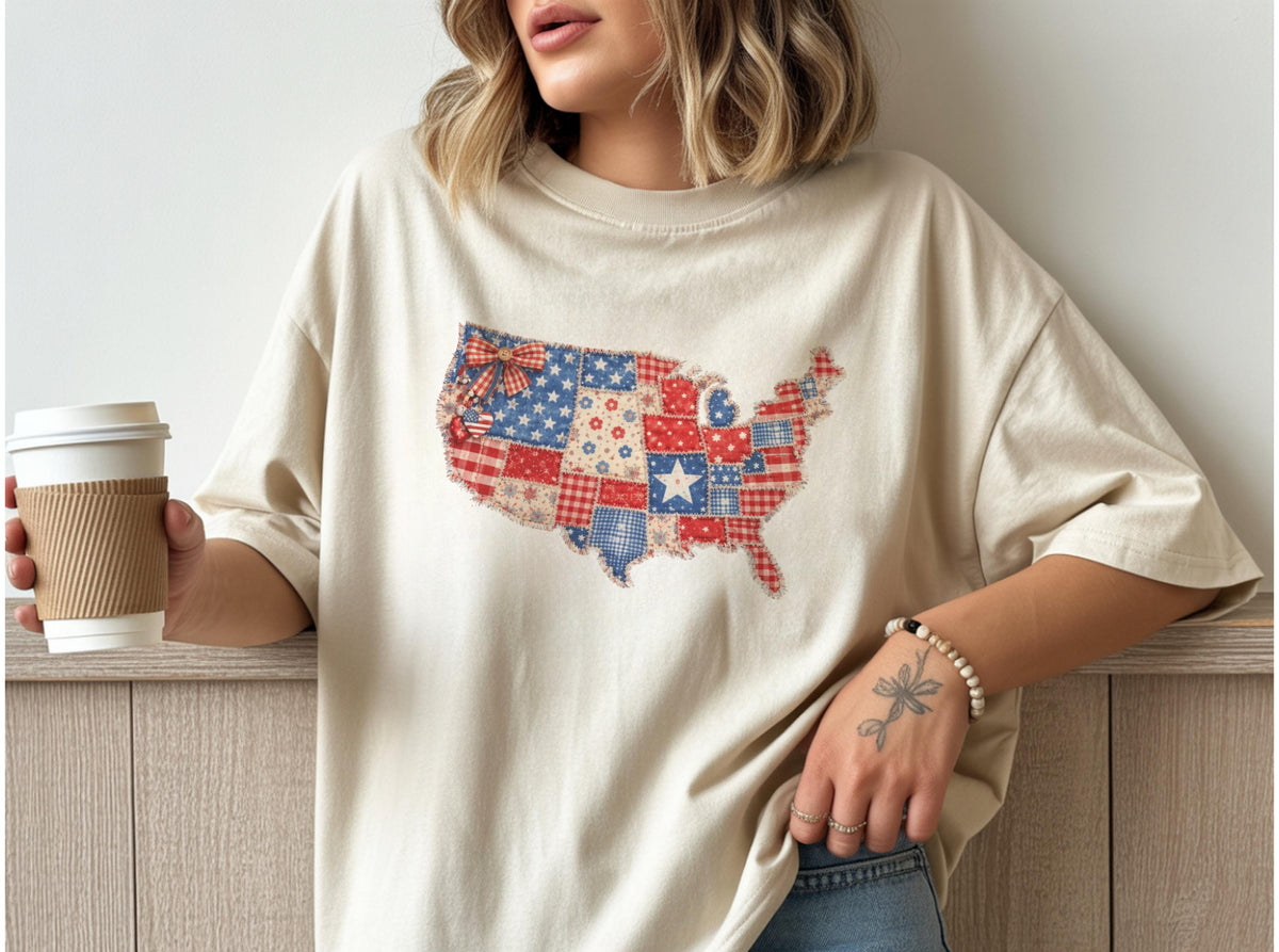 May include: A cream-colored t-shirt featuring a patchwork design of the United States map. The map is composed of red, white, and blue fabric squares, stars, and stripes. The shirt has short sleeves and a relaxed fit.