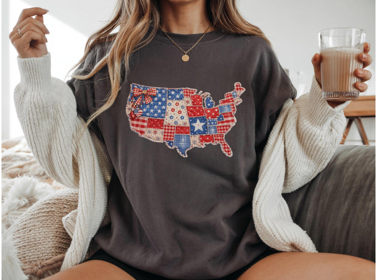 May include: Dark gray sweatshirt featuring a patchwork design of the United States map in red, white, and blue. The map design includes stars, stripes, and floral patterns. The sweatshirt is worn with a cream-colored cardigan.