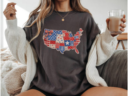May include: Dark gray sweatshirt featuring a patchwork design of the United States map in red, white, and blue. The map design includes stars, stripes, and floral patterns. The sweatshirt is worn with a cream-colored cardigan.
