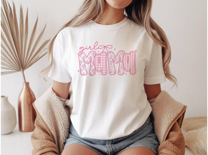 May include: White t-shirt with the text "girl Mama" in pink. The word "Mama" is designed with floral, gingham, and striped patterns. A pink bow is above the word "girl".