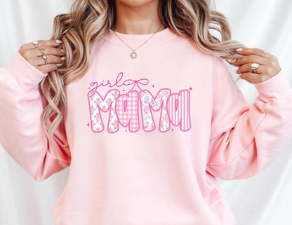 May include: A light pink sweatshirt with the words "girl Mama" in a decorative font. The word "Mama" is in a bubble letter style with a floral and gingham pattern. The sweatshirt is a solid light pink color.